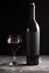 A bottle of red expensive wine with a black blank label on a dark background with a glass of red wine.