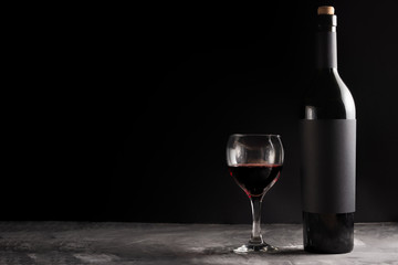 A bottle of red expensive wine with a black blank label on a dark background with a glass of red wine. Wine bottle mockup with space for text.