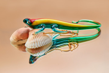 Colored glass lizard on shells with reflection on beige-sand background