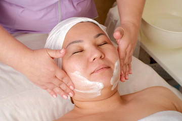 Obraz na płótnie Canvas A woman of Asian appearance with closed eyes on a face skin cleaning procedure in a beauty salon. The hands of a cosmetologist applied soap suds on the patient's cheeks.