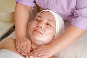 Obraz na płótnie Canvas The hands of a professional cosmetologist put soap suds on the female neck of the patient. A woman of Asian appearance with closed eyes on a face skin cleaning procedure in a beauty salon. Close-up.