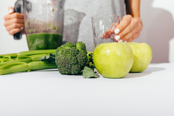 Young woman in gray t-shirt holding blender with healthy green smoothie and empty glass