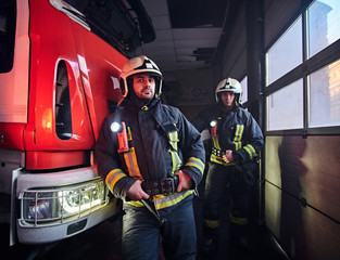 Two firemen wearing protective uniform standing next to a fire engine in a garage of a fire department.