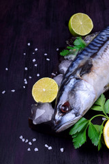 Fresh mackerel on a black chopping board surrounded by ice, lime, lemon, parsley and spices