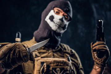 A terrorist in a military uniform and a skull balaclava holding a pistol and a knife and looks at...