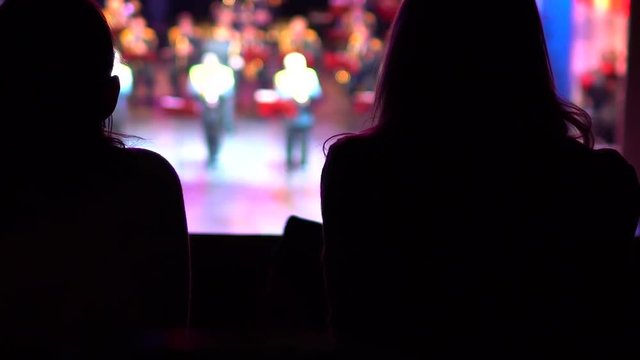 Silhouettes of two women who sit in the opera house or concert hall and watch the performance on a bright bright stage.