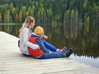 Young beautiful European lady and her son are sitting on the wooden quay near the river. They are enjoying their holidays and serenity round. The boy is pointing somewhere on the other side.