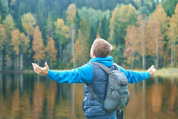 Young man with a backpack on his shoulders having a rest in a picturesque place near the river bank, he is enjoying the view, his hand are open wide...