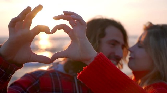 Happy young couple making heart shape with hands and kissing at the sunset on a lonely beach