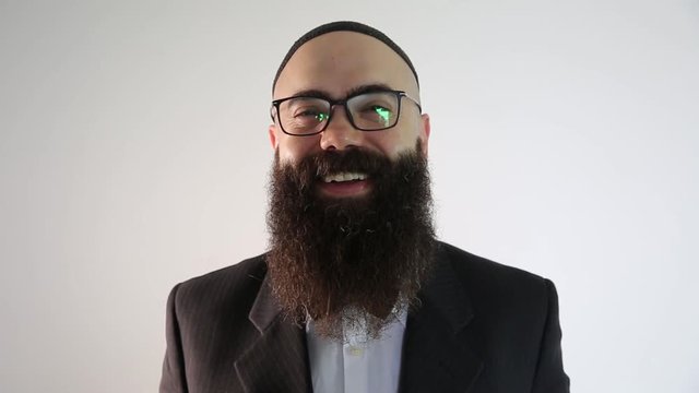 A bearded man with quip and a traditional Jewish clothing. He smiles looking at the camera.