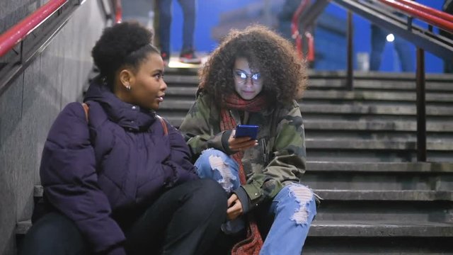 Two young multiethnic women sitting in the city night using smart phone talking– social network, browsing, wireless communication concept