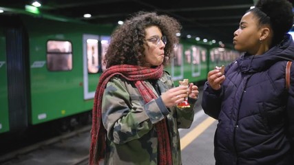 Two young multiethnic women in the underground eating snack – unhealthy eating, satisfaction, sweet food concept