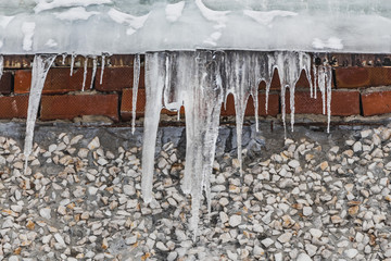 A group of sharp white transparent icicles is hanging down from the gray roof with ice of a building from red bricks and white stones