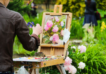 Painter artist draws a bouquet of peonies in the Park. Artist painting on easel in the garden. Girl paints flowers with brush, spring landscape. Female painter seen from behind.Art, creativity concept