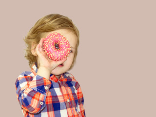 Little happy cute boy is eating donut on brown background wall. Child is having fun with donut. Tasty food for playing kids. Funny time  with sweet food. Bright baby boy in a plaid shirt.
