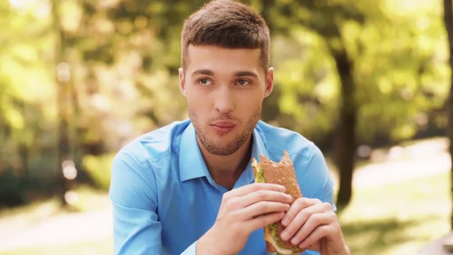 Handsome young businessman eating very tasty sandwich in a sunny park. Young man in a blue shirt eating lunch out of office. Portrait shot