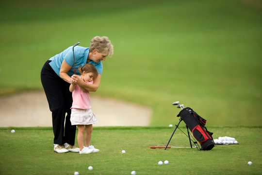 Senior woman teaching her young granddaughter how to swing a golf club on a golf course.