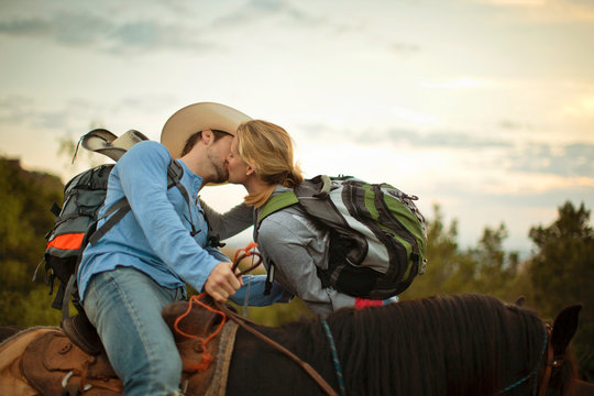Affectionate young couple take a break from horse trekking to share a horseback kiss.