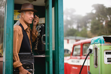 Mature man wearing a hat while listening on the phone in a public phone booth holding a typewriter.