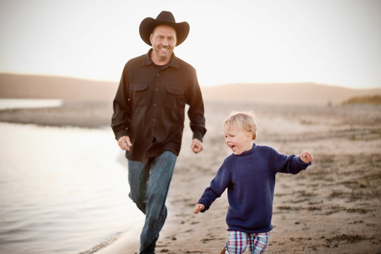 Middle aged man wearing a cowboy hat plays with his toddler son on the beach. 