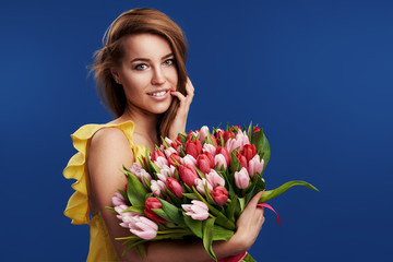 Perfect gift. Wonderful flowers on Womens Day. Tender smiling brunet woman poses with colorful tulips, looks happily, isolated over blue studio background. Space for text