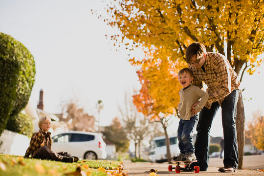 Father helping son balance on skateboard while brother watches from lawn.