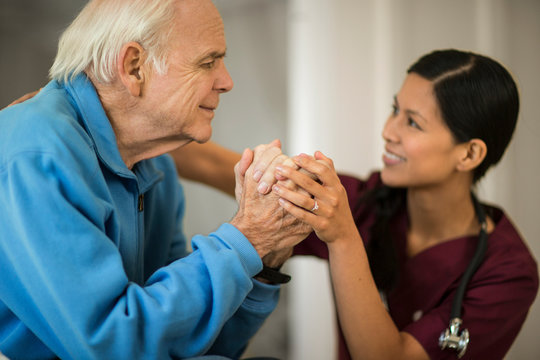 Smiling female nurse holding the clasped hands of an elderly male patient as they have a conversation.