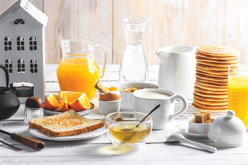 Healthy breakfast eating concept, various morning food - pancakes, soft-boiled egg, toast, oatmeal, granola, fruit, coffee, tea, orange juice, milk on white wooden table