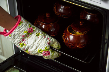 Woman taking out of oven stewed or baked potatoes in brown ceramic pots.