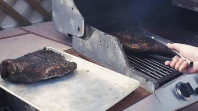 Man pulls flame grilled beef off a barbecue grill. Tracking shot of using bbq tongs to let the delicious smoked meat rest before serving it at a summer picnic. Action shot of grilling competitions.