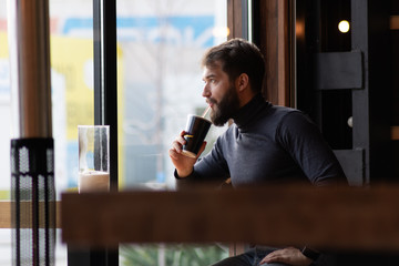 handsome bearded guy drinks a drink from a straw near the window in a cafe.  attractive young stylish man sitting in a cafe and looking out the window. Business, freelance