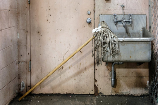 Dirty old mop , leaning on a grimy ,filthy, stainless shop sink ,in a janitorial cleaning supply room.