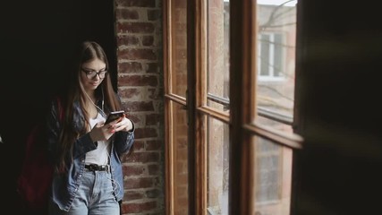 Young girl standing by the window and texting while listening to music