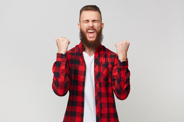 Happy bearded man in checkered shirt clenching fists like winner with eyes closed in pleasure, shouts about his victory celebrates his win, isolated over white background