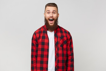Fototapeta premium Excited surprised attractive young bearded guy in checkered shirt, opened mouth because of amazement, with mustache fasion hairstyle, isolated on white background