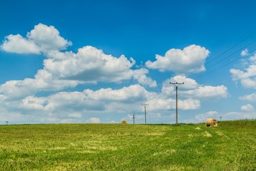 Fototapeta na wymiar Rural countryside with harvested agriculture field, straw bales, electricity poles and lines on horizon, sunny summer day, bright blue sky with white clouds