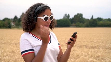 Slow motion tracking video clip of beautiful mixed race African American girl teenager young woman wearing white sunglasses walking listening to music on wireless headphones at sunset or sunrise