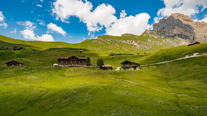 Group of beautiful mountain huts on a gentle slope of the Dolomites with a rocky rocky peak in the background. Blue sky with big white clouds.