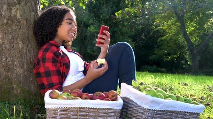 Beautiful African American mixed race teenage girl young woman sitting leaning against a tree in an orchard eating an apple using her mobile cell smart phone for social media