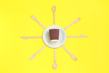 Eco-friendly disposable utensils made of bamboo wood that draped spoons, fork, knives, around white paper plate on yellow background.