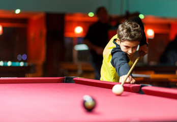 Cute boy in yellow t shirt plays billiard or pool in club. Young Kid learns to play snooker. Boy with billiard cue strikes the ball on table. Active Leisure, sport, hobby concept