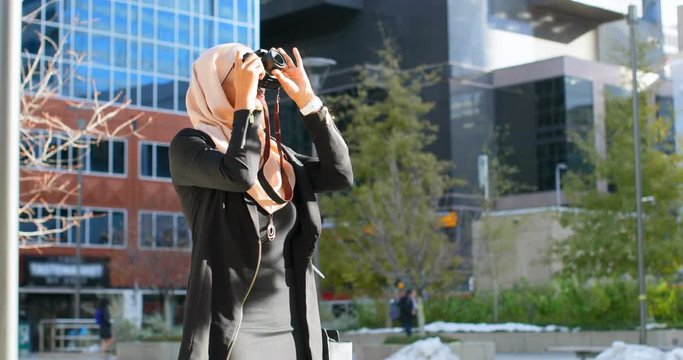 Front view of young Asian woman in hijab clicking photos with digital camera in the city 4k