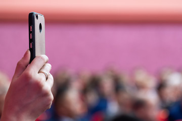 Smartphone in a female hand. Auditorium. Blurred pink background. Photo or video on a mobile phone. Live broadcast with the help of a modern gadget.
