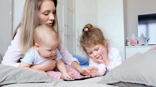 Positive family mother, daughter and son are cheerfully watching a tablet with children's films sitting in a cozy bedroom. Concept of care and education for children