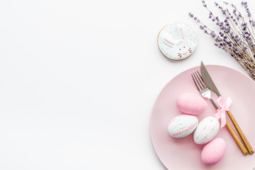 Easter dining table in pastel colors. Plate, cutlery, painted eggs, gingerbread and lavender branches on white background top view copy space