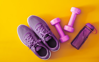 Top view of purple and violet sport shoes, dumbbells and bottle of water on yellow background....