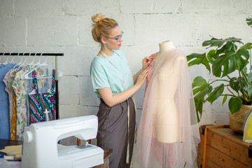 Young caucasian seamstress inserts needles into a lace wedding dress dressed on dummy. Creation of...
