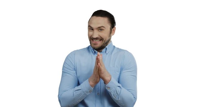 Handsome cheerful young man in the blue shirt standing on the white screen background, smiling archly while rubbing his hands.