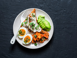 Cream cheese toast, avocado, boiled egg, baked sweet potatoes - delicious healthy breakfast or snack on a dark background, top view