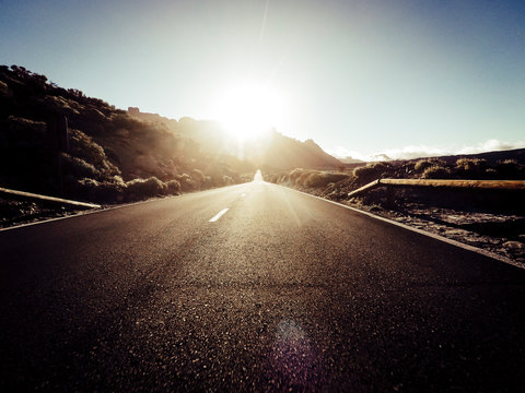 Long Way Road At The Mountain With Sun In Front And Sunlight Effect - Ground Point Of View With Black Asphalt And White Lines - Driving And Travel Concept